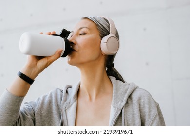 Asian Mature Woman In Headphones Drinking Water During Workout Outdoors