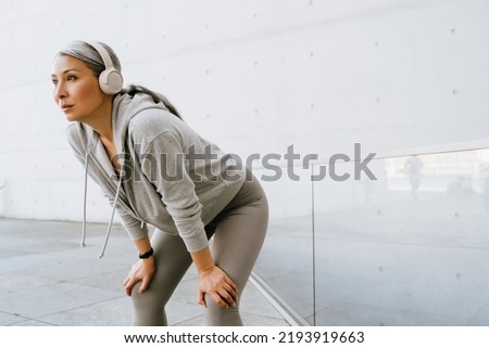 Asian mature woman in headphones standing during workout outdoors