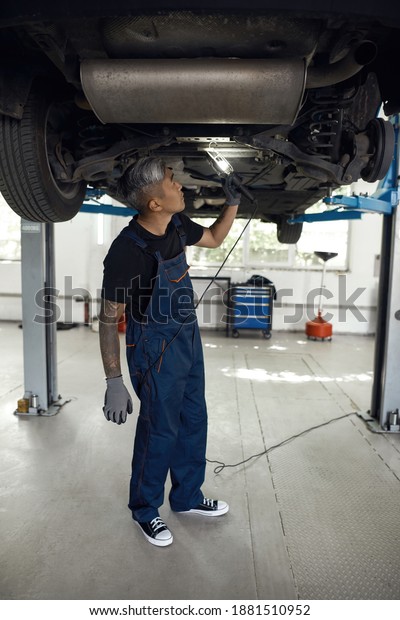 Asian mechanic in car service station\
uniform standing under auto raised on lift using flashlight for\
diagnostic, vertical shot. Car repairing\
concept