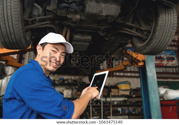 Asian mechanic looking under the car to\
repair the engine with tablet in hand, japanese mechanic portrait\
style, mechanic maintenance working under\
car