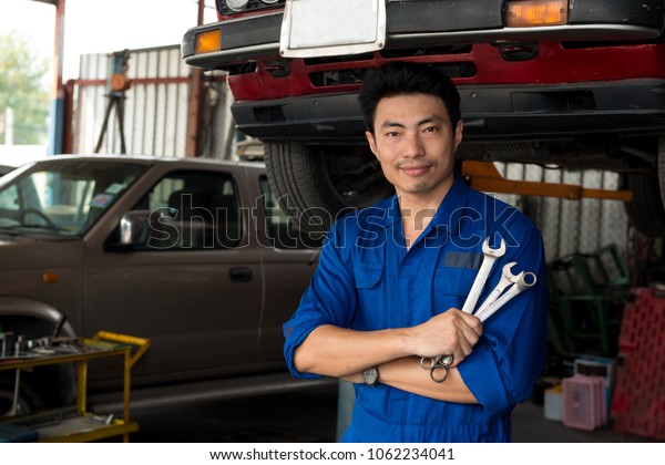 Asian mechanic looking under the car\
pose looks at camera and hold tools in hand, japanese mechanic\
portrait style, mechanic maintenance working under\
car
