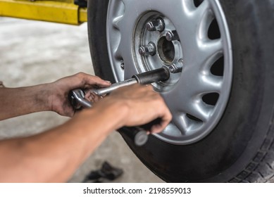 An Asian Mechanic Loosens And Removes The Lug Nuts From The Tire Of A Serviced Car With A Ratchet Lug Wrench. Removing A Wheel Manually. At An Auto Repair Shop.