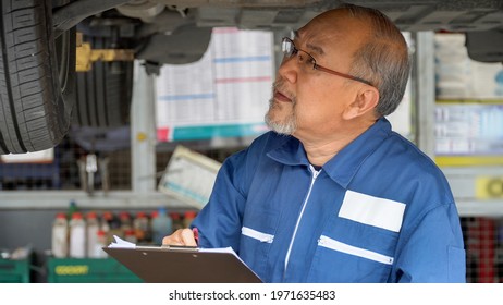 Asian Mechanic Senior Man Holding Clip Board Checking List To Brake ,tyre, Undercarriage Of Car In Workshop At Auto Car Repair Service Center With Lift. Car Engineer Old Man Inspection Vehicle Details