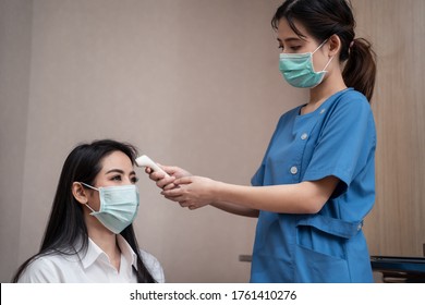 Asian Medic Nurse Measure And Check Patient Temperature From Infrared Thermometer Scan At Forehead In Hospital Examination Room. Women Wear Face Surgical Mask To Prevent Covid Virus Infection.