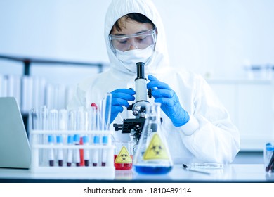 Asian Medical Scientist In Protective White Jumpsuit And Glasses And Face Mask Diagnosis Virus With Microscope In Clinical Laboratory