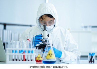 Asian Medical Scientist In Protective White Jumpsuit And Glasses And Face Mask Diagnosis Virus With Microscope In Clinical Laboratory