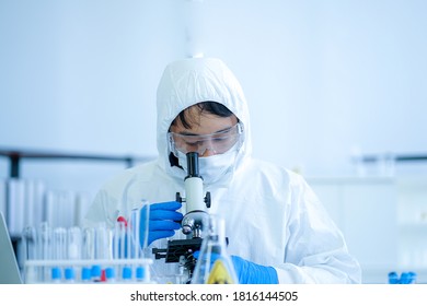 Asian Medical Scientist In Protective White Jumpsuit And Glasses And Face Mask Diagnosis Virus With Microscope In Clinical Laboratory