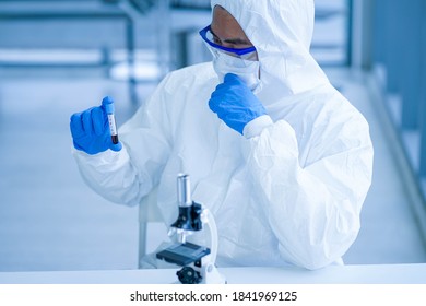 Asian Medical Scientist Wearing Protective White Jumpsuit And Face Mask With Glasses Testing Sample With Microscope In Clinical Laboratory