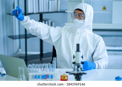 Asian Medical Scientist Wearing Protective White Jumpsuit And Face Mask With Glasses Testing Sample With Microscope In Clinical Laboratory