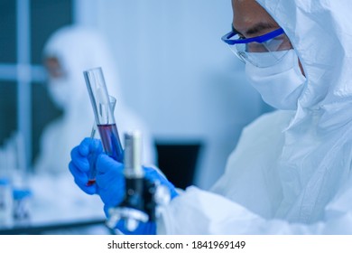 Asian Medical Scientist Wearing Protective White Jumpsuit And Face Mask With Glasses Testing Sample With Microscope In Clinical Laboratory