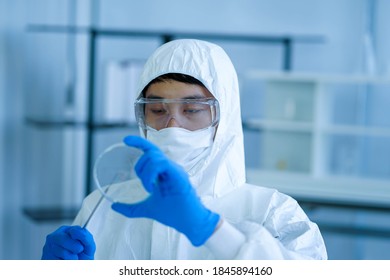 Asian Medical Scientist Wearing Protective White Jumpsuit And Face Mask With Glasses Testing Sample With Microscope In Clinical Laboratory