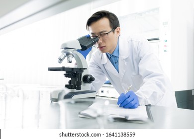 Asian Medical Scientist Wearing White Coat Examining Specimen Using Microscope, Horizontal Portrait