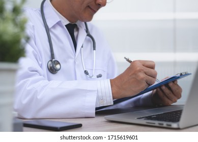 Asian Middle Age Doctor Review Patient Information, Hand Writing Prescription, Filling On Paper Document And Working On Laptop Computer In Medical Office In Hospital, Close Up