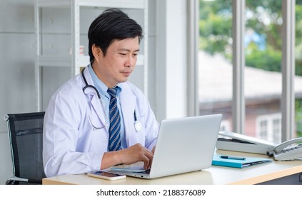 Asian Middle Aged Male Professional Successful Doctor Practitioner In White Lab Coat With Stethoscope Sitting Working At Workstation Desk Typing Laptop Notebook Computer In Clinic Hospital Office.