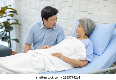 Asian Middle Aged Male Son Visiting Supporting Comforting Holding Hands Old Senior Sick Unhealthy Pensioner Mother Patient In Blue Hospital Uniform Laying Down Receiving Saline Solution Drip On Bed.