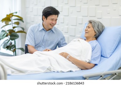 Asian Middle Aged Male Son Visiting Supporting Comforting Holding Hands Old Senior Sick Unhealthy Pensioner Mother Patient In Blue Hospital Uniform Laying Down Receiving Saline Solution Drip On Bed.