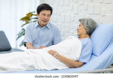 Asian Middle Aged Male Son Visiting Supporting Comforting Holding Hands Old Senior Sick Unhealthy Pensioner Mother Patient In Blue Hospital Uniform Laying Down Receiving Saline Solution Drip On Bed.