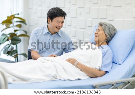 Asian middle aged male son visiting supporting comforting holding hands old senior sick unhealthy pensioner mother patient in blue hospital uniform laying down receiving saline solution drip on bed.
