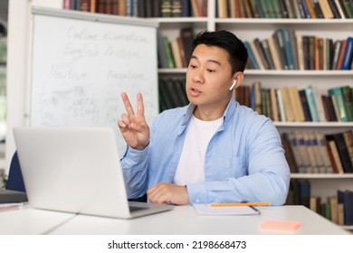 Asian Middle Aged Male Teacher Making Video Call Using Laptop Computer Teaching And Working Online Sitting At Desk In Library Indoors. E-Teaching And Modern Communication Concept