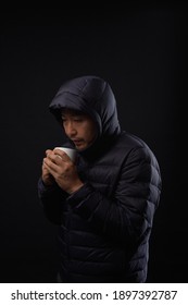 Asian middle aged man in anorak  holding cup drinking hot water to get warm on black background. Homeless concept.