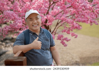 Asian middle-aged man in white cap and has backpack standing and thumping up in front of sakura flower tree, soft focus, happiness of middle-aged people and a single man around the world concept. - Powered by Shutterstock