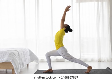 Asian Middle-aged Woman Practice Yoga In The Bedroom. Warrior Posture. Stand One Knee Forward The Other Leg Stretched Straight Back Hold The Two Arms Above The Head On Top.