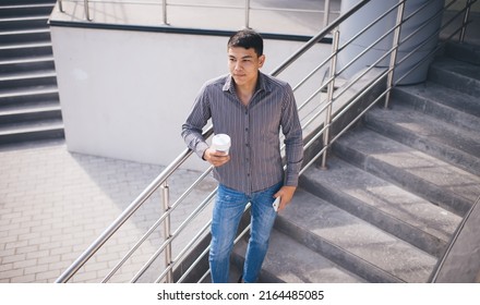 Asian Millennial Guy With Mobile Phone And Coffee Standing On Stairs Near Office Building. Concept Of Rest And Break At Work. Idea Of Modern Successful Man. Young Male Person Looking Away. Sunny Day