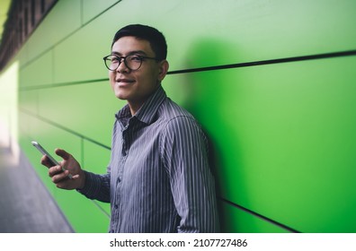 Asian Millennial Guy With Smartphone Looking At Camera On Green Wall Background. Concept Of Remote And Freelance Work. Idea Of Modern Successful Man. Young Smiling Male Person In Glasses