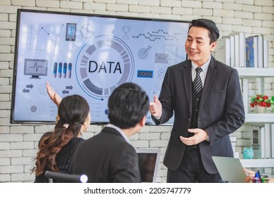 Asian millennial professional successful businessman in black formal suit standing presenting showing company data on big monitor screen with male and female colleagues in meeting room workstation. - Shutterstock ID 2205747779