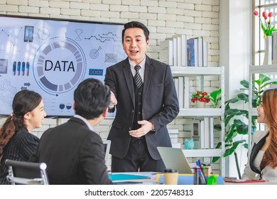 Asian millennial professional successful businessman in black formal suit standing presenting showing company data on big monitor screen with male and female colleagues in meeting room workstation. - Shutterstock ID 2205748313