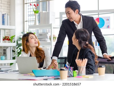 Asian Millennial Professional Successful Male Businessman Mentor In Black Formal Suit Standing Helping Two Female Employee Sitting Working With Laptop Computer At Workstation In Company Meeting Room.