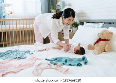 Asian Millennial Woman Interacting With Her Daughter Before Changing Clothes For Her. Pleasant Ambience Japanese Mom Playing With Her Baby Lying On Bed Scatter With Baby Clothes