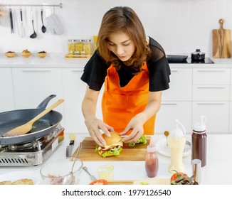 Asian Modern Pretty Mom Wearing Orange Kitchen Apron Carefully Arrange  Hamburger At Home As Lovely Breakfast For Kid And Beloved Family.