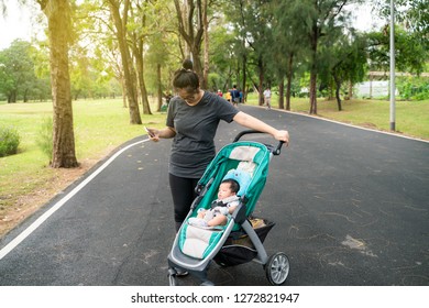 Asian Mom With Baby Boy In Stroller Walking In Green Park, Family In Garden