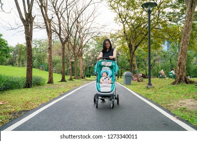 Asian Mom With Baby Boy In Stroller Walking In Green Park, Family In Garden