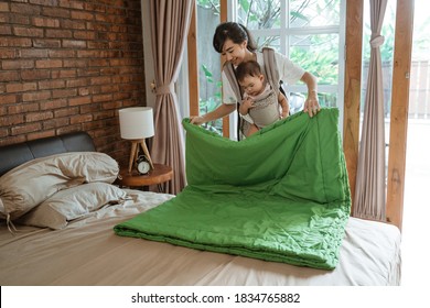 Asian Mom Carries The Baby Cleaning And Arranges Blanket On The Bed In The Bedroom,House Cleaning Concept