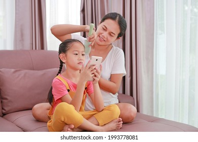 Asian Mom Combing Hair For Her Daughter While Playing Smartphone On Sofa In Living Room At Home