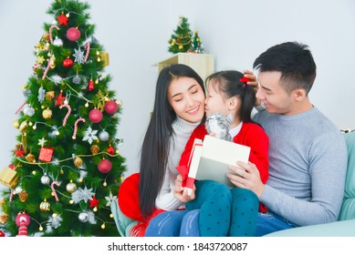 Asian Mom And Dad Opening Gift Box With Her Daughter. Near The Christmas Tree At Home. Little Girl Kissing Her Mother,thanks For Present.