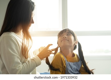 Asian Mom Making Hair Braid For Daughter While Sitting On Bed At Home.