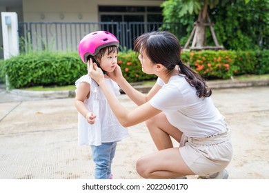 Asian Mom Put On Sport Helmet To Little Kid For Safety Before Exercise. Beautiful Loving Mother Wear Protective Hard Hat To Young Girl Daughter Before Ride Bicycle For Workout On Street In The Morning