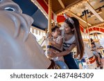 Asian mother and adorable kid daughter riding carousel at theme park. Happy family, attractive woman and little girl feeling happy and relax while traveling for holiday vacation trip at amusement park