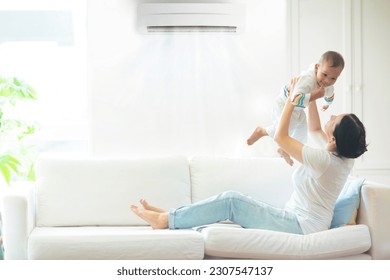 Asian mother and child with air conditioner remote control. Comfortable temperature at family home. Cooling device. Mom and baby boy on couch under cold breeze. Air conditioning on hot summer day. - Powered by Shutterstock