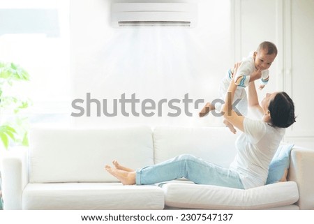 Asian mother and child with air conditioner remote control. Comfortable temperature at family home. Cooling device. Mom and baby boy on couch under cold breeze. Air conditioning on hot summer day.