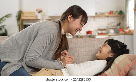 Asian Mother And Daughter Having Fun Playing Tickle Together On Living Room Couch At Home. The Girl Pushes Her Mom Away While Laughing With Excitement