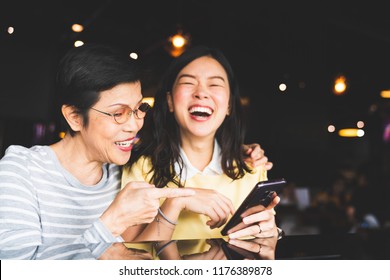 Asian mother and daughter laughing and smiling on a selfie or photo album, using smartphone together at restaurant or cafe, with copy space. Family love, holiday activity, or modern lifestyle concept