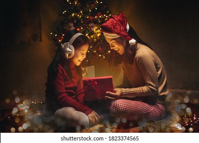 Asian Mother And Daughter Open Christmas Lighting Present Gift Box Front Of Xmas Tree. Happy Asia Mother With Kid Girl In Magic Night. Family Merry Christmas And Newyear Holiday Concept.