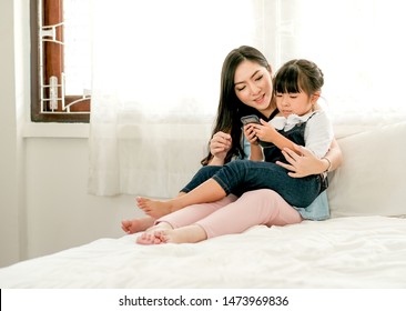 Asian Mother And Daughter Stay On White Bed And Look To The Mobile Phone And Enjoy Together. 