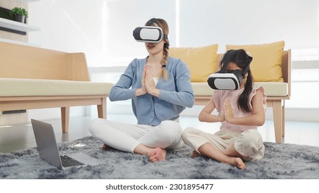 Asian mother and daughter wearing glasses of technology virtual reality headset doing yoga exercises in lotus position sitting on floor in living room. VR metaverse technology concept - Powered by Shutterstock