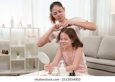 Asian mother gently fixing daughter's hair. Smiling child holding mirror, surrounded by beauty products at home. Warm and caring atmosphere during beauty routine. - Powered by Shutterstock