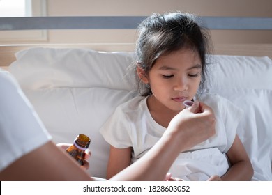 Asian Mother Giving Cough Syrup Medicine On A Spoon To Her Daughter In Bed, Sick Child And Health Care Concept.